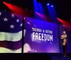 A person stands on stage beside a large screen displaying the American flag and the words Cherish Defend Freedom