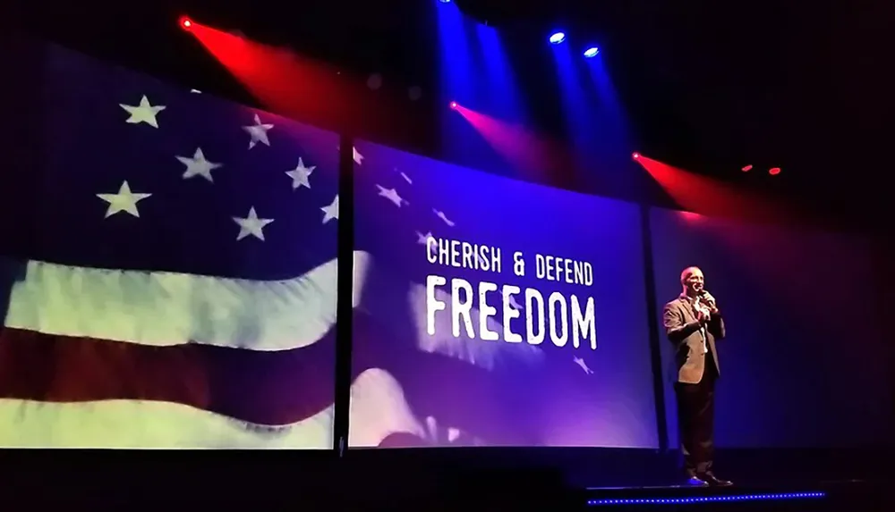 A person is speaking on stage with a backdrop displaying the American flag and the words Cherish  Defend Freedom under dramatic red and blue lighting
