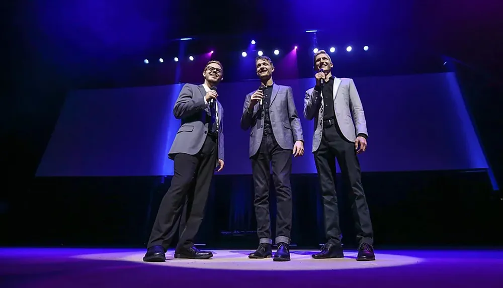 Three men in suits are standing on stage with microphones under bright purple and blue stage lights