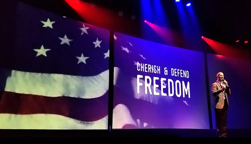 A man in a suit speaks on stage with large screens behind him displaying the American flag and the words Cherish  Defend Freedom