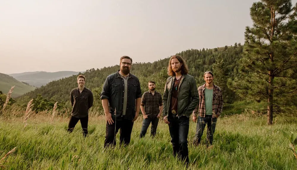Five men are standing in a grassy field with hills and trees in the background