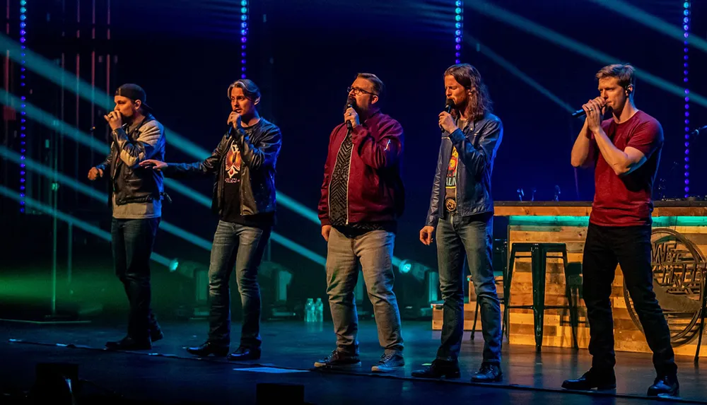 Five men are performing on stage with microphones under colorful stage lights