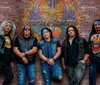 Five men dressed in rock-style attire pose in front of a brick wall with a colorful BON JOUR mural featuring a heart and wings design