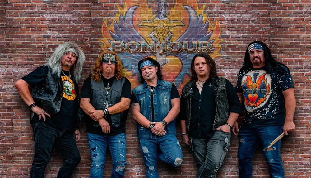 Five men dressed in rock-style attire pose in front of a brick wall with a colorful BON JOUR mural featuring a heart and wings design