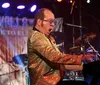A performer in a sparkly outfit and colorful glasses is playing the piano and smiling on stage under bright lights and smoke effects