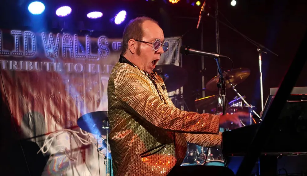 A musician wearing a sparkly jacket is energetically playing the keyboard on stage with colorful lights in the background