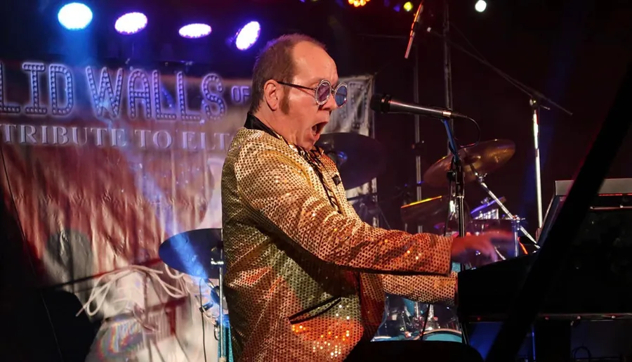 A musician wearing a sparkly jacket is energetically playing the keyboard on stage with colorful lights in the background.