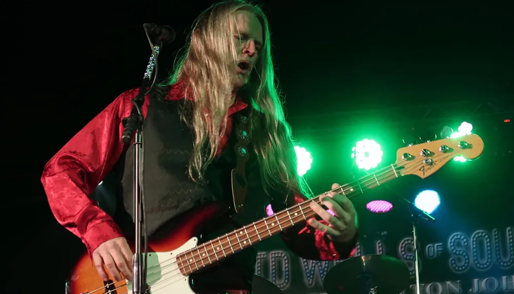A musician with long hair passionately plays a bass guitar on stage under colorful lights