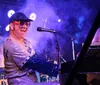 A performer in a sparkly outfit and colorful glasses is playing the piano and smiling on stage under bright lights and smoke effects