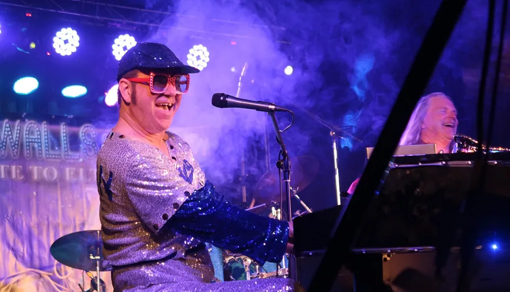 A performer in a sparkly outfit and colorful glasses is playing the piano and smiling on stage under bright lights and smoke effects