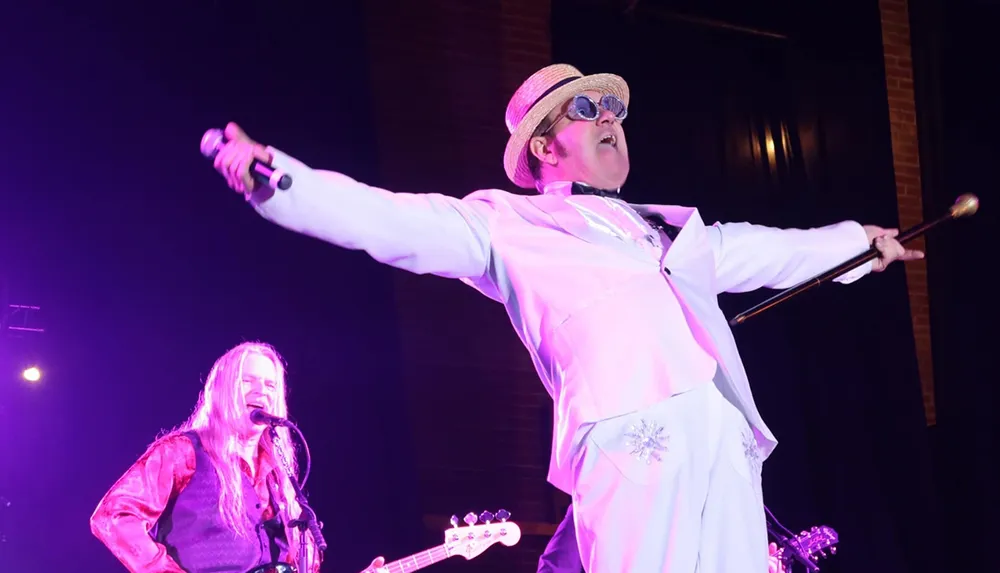 A performer in a white suit and straw hat holding a microphone and cane energetically sings on stage with another musician playing guitar in the background