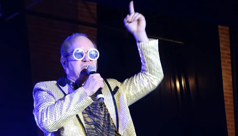 A person wearing oversized round glasses and a shiny jacket is speaking into a microphone gesturing with one finger pointed upwards