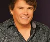 A person in a patterned blue shirt is smiling against a wooden background