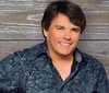 A person in a patterned blue shirt is smiling against a wooden background