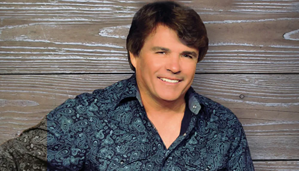 A person in a patterned blue shirt is smiling against a wooden background