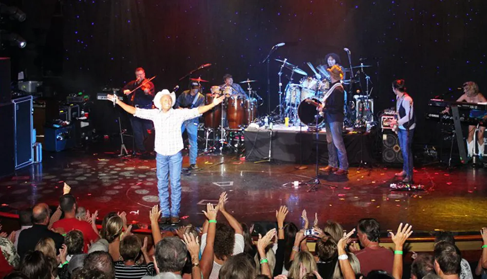 A musician in a cowboy hat stands with arms open on stage surrounded by a band performing for an enthusiastic crowd
