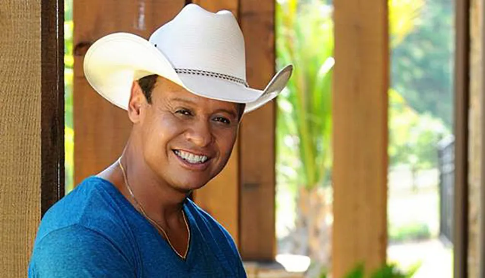 A smiling person in a white cowboy hat and blue shirt is standing outdoors near a wooden structure
