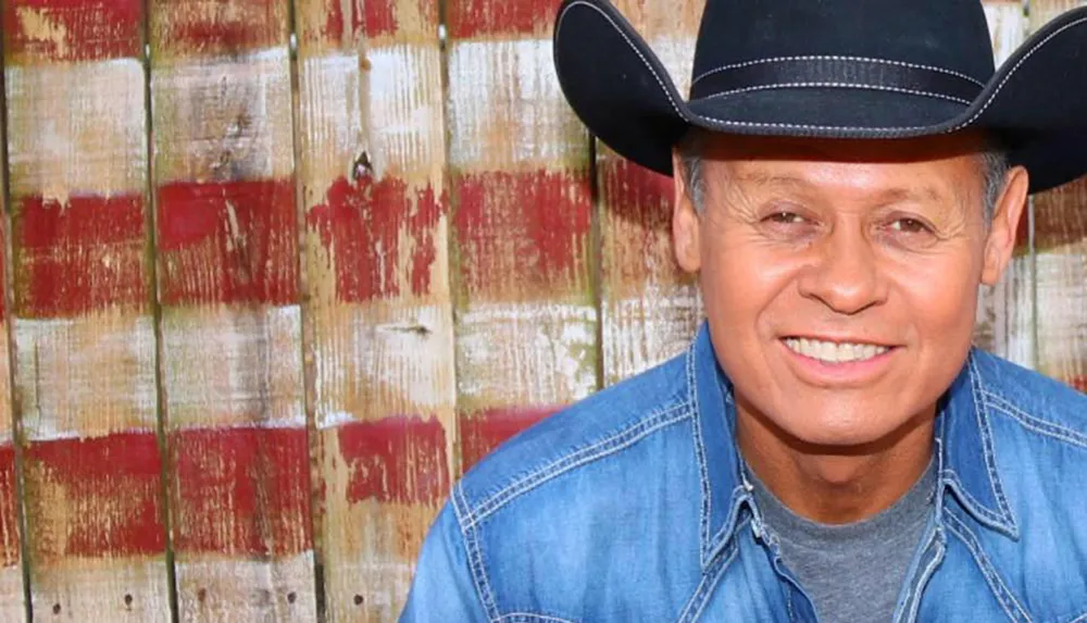 A person wearing a black cowboy hat and blue denim shirt smiles in front of a rustic wooden background with red and white paint