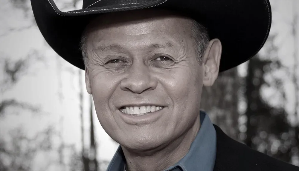 A smiling person is wearing a cowboy hat and a dark-colored jacket with a blurred outdoor background