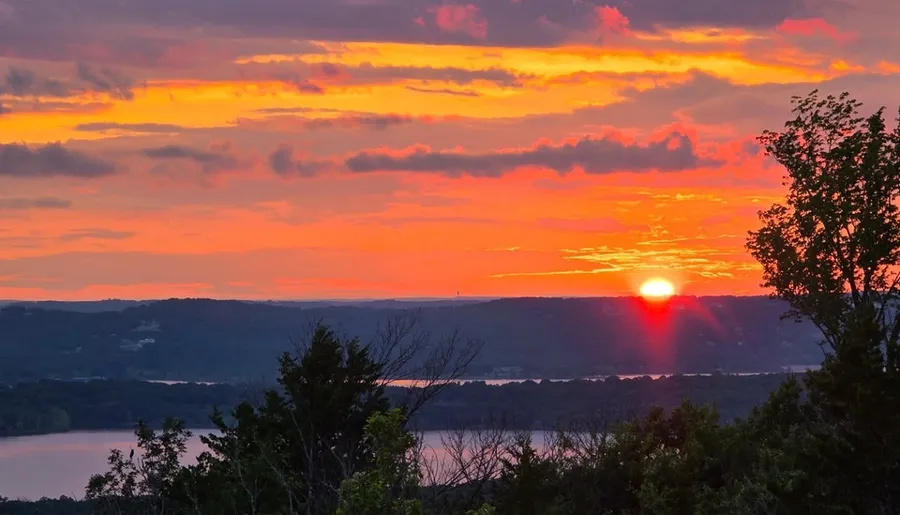 A vibrant sunset illuminates the sky with hues of orange and pink above a serene landscape featuring a lake and silhouetted trees.