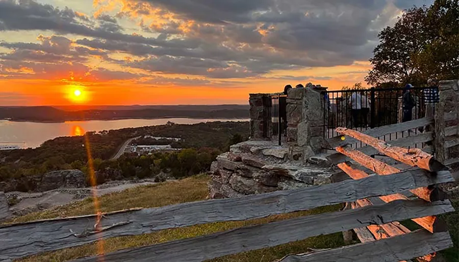 A scenic sunset view with the sun reflecting over a body of water and silhouetted people near a stone structure and wooden fence.