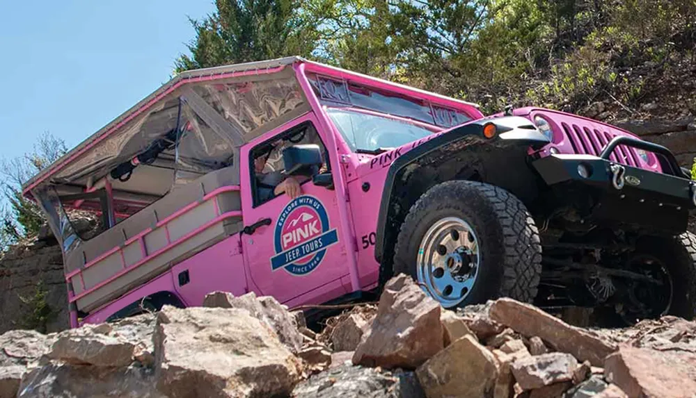 A pink Jeep is navigating a rocky off-road terrain amidst trees and rough slopes