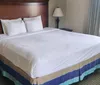 The image shows a neatly made bed with white linens and two pillows flanked by a wooden headboard a nightstand and a lamp in a well-lit room