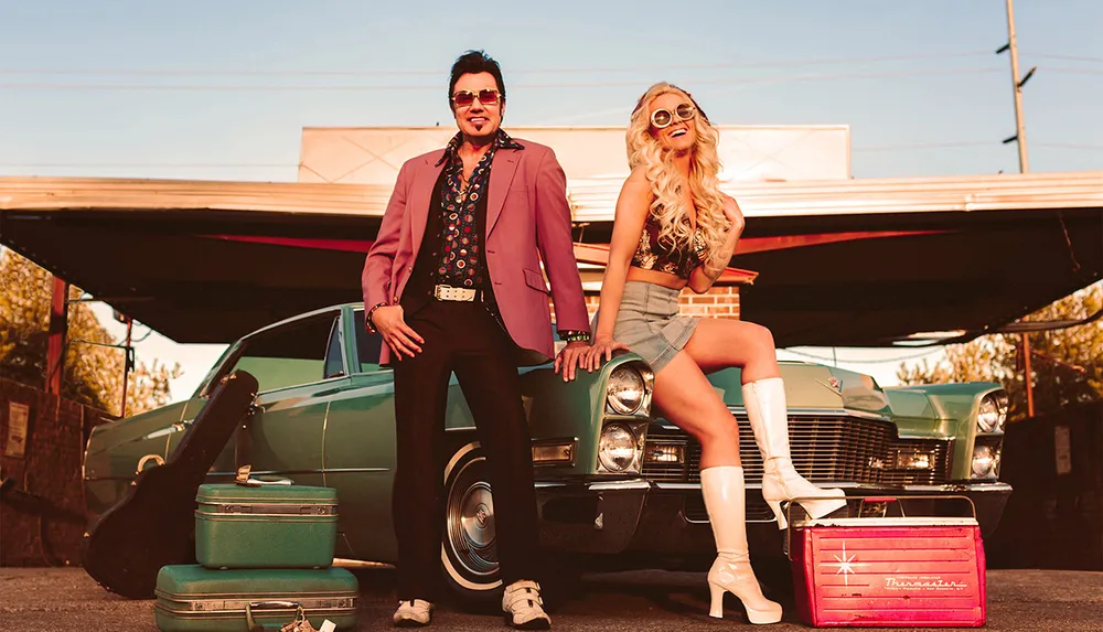 A man and a woman dressed in retro attire pose confidently in front of a vintage car with old-fashioned luggage nearby