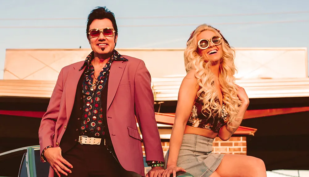 A man and woman both wearing stylish retro outfits and sunglasses pose confidently outdoors