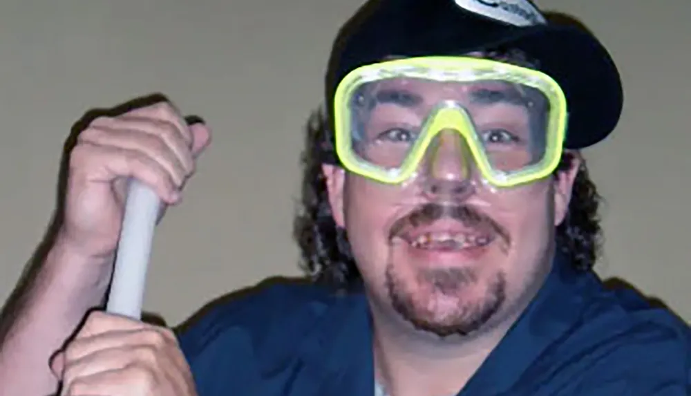 A person wearing a backwards cap and bright goggles is smiling while holding a stick