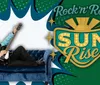 A man in a blue and black outfit is reclining on a blue sofa with a guitar next to a vibrant RocknRoll SUN Rise sign