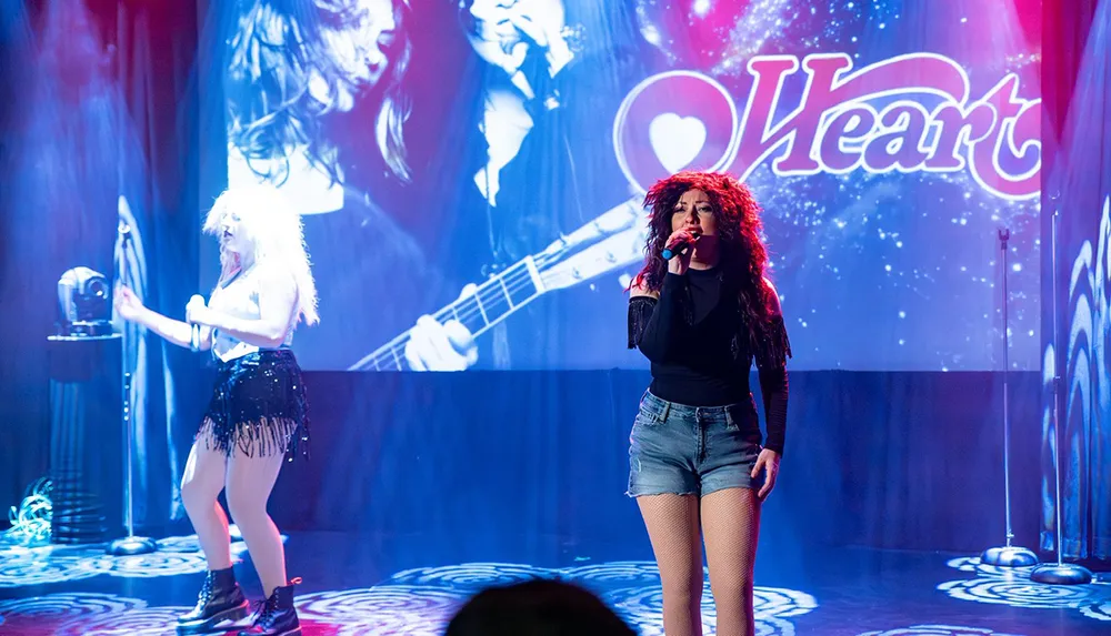 Two performers are on stage with vibrant lighting and a large projection in the background displaying the word Heart