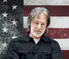 A man with gray hair and a beard stands in front of a weathered American flag