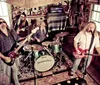 A four-member band is energetically performing with guitars and drums in a room filled with music equipment and posters