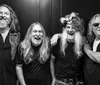 The black and white image shows four men with long hair and beards smiling and posing together with one wearing a fur hat and another wearing sunglasses and holding a cigar
