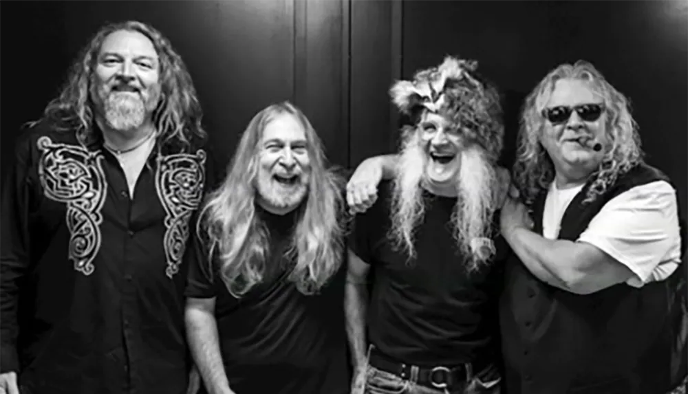 The black and white image shows four men with long hair and beards smiling and posing together with one wearing a fur hat and another wearing sunglasses and holding a cigar