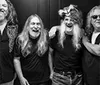 The black and white image shows four men with long hair and beards smiling and posing together with one wearing a fur hat and another wearing sunglasses and holding a cigar