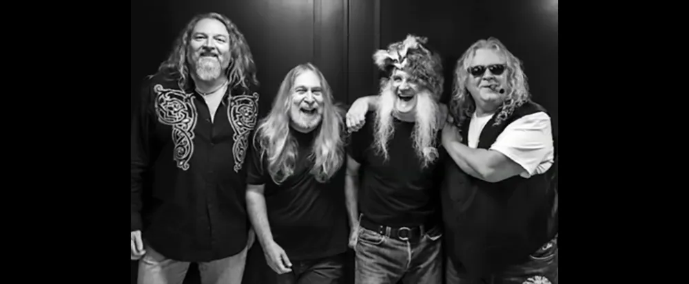 Four men with long hair and beards are smiling and posing together with one wearing a unique furry hat
