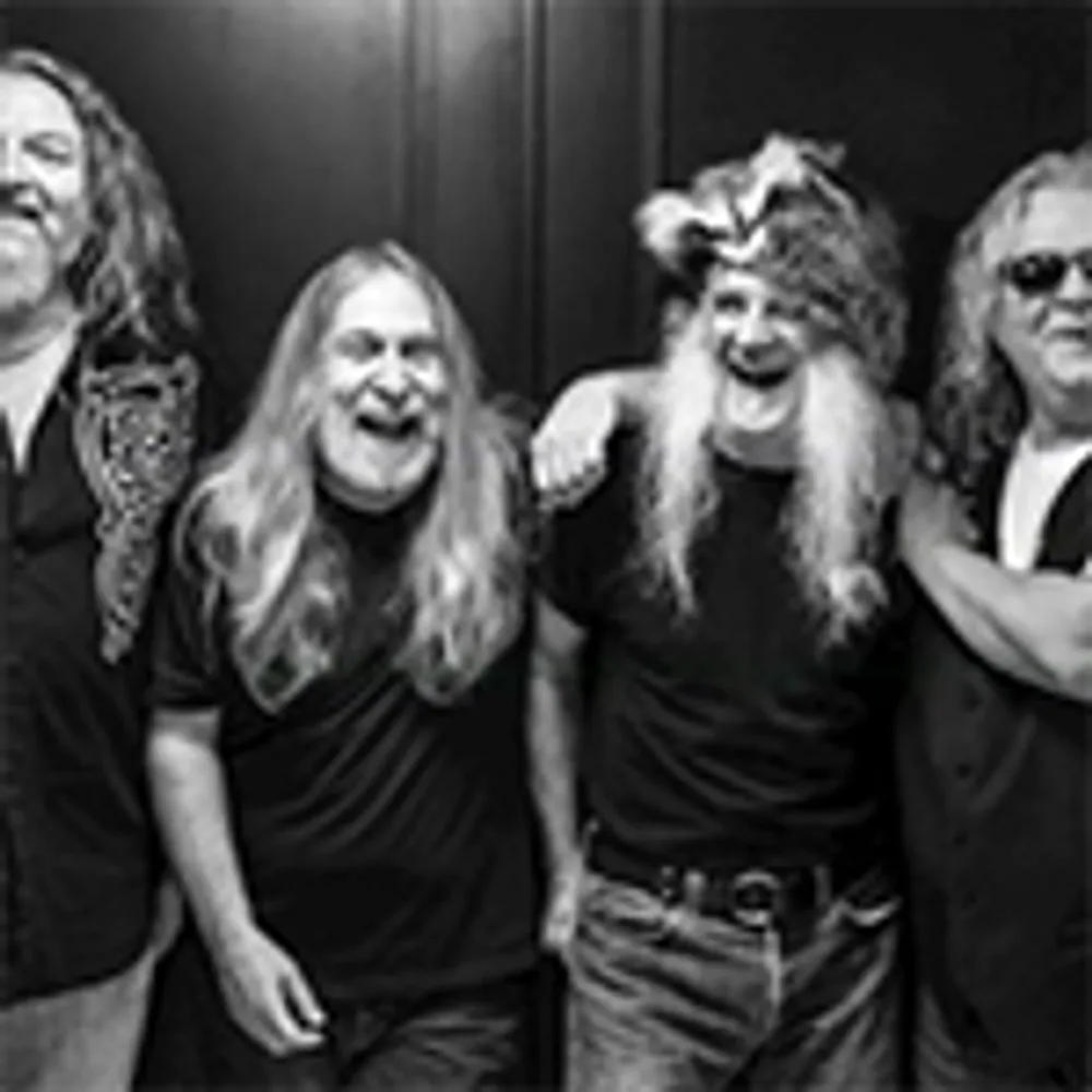 A black-and-white photo of four smiling men with long hair standing together and posing for the camera