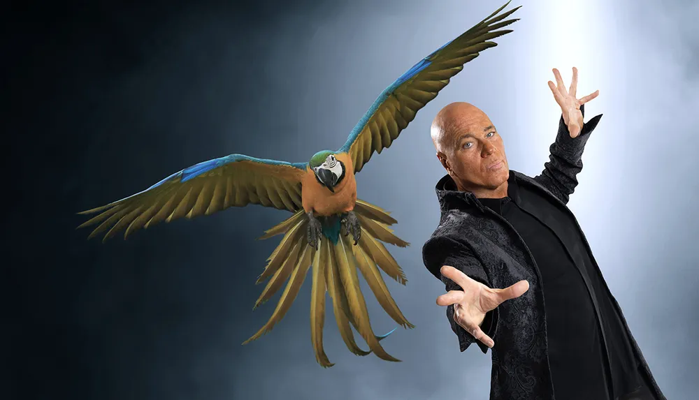 A man in a dark patterned jacket is striking a dramatic pose beside a colorful parrot in flight against a smoky background