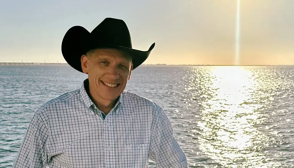 A person wearing a cowboy hat is standing in front of a body of water during a bright sunny day