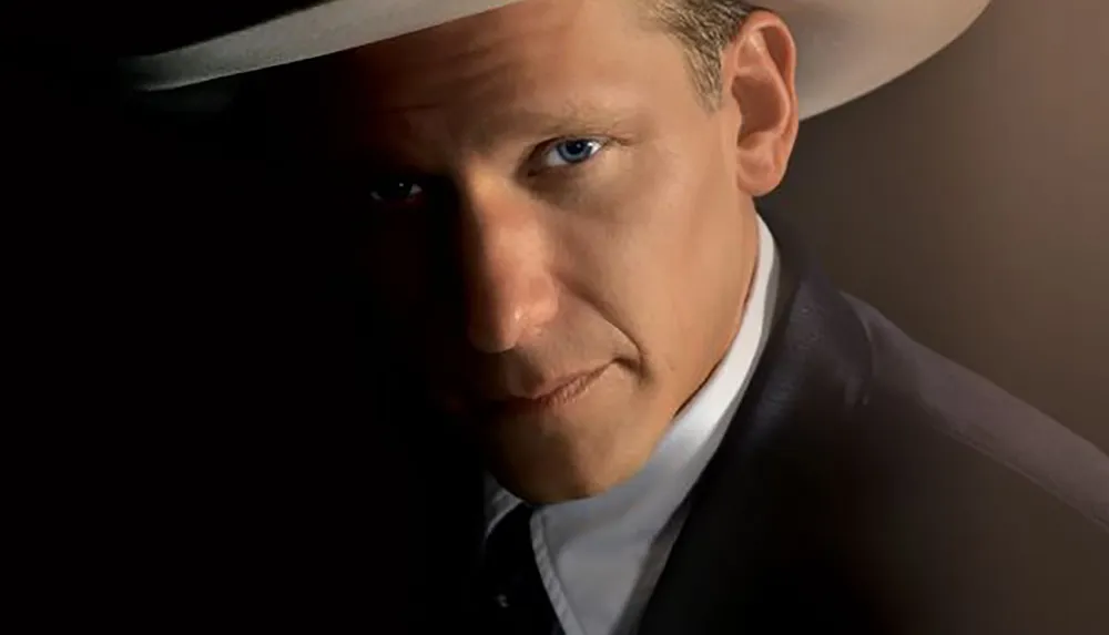 A person wearing a white cowboy hat looks directly at the camera partially illuminated in a dimly lit setting
