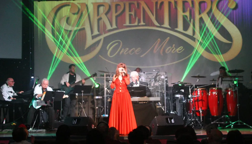A band is performing on stage with a woman in a red dress singing under green stage lights with Carpenters Once More displayed in the background