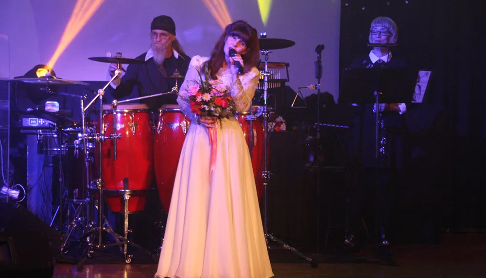 A woman in a long dress sings into a microphone while holding a bouquet with musicians playing drums and a saxophone in the background