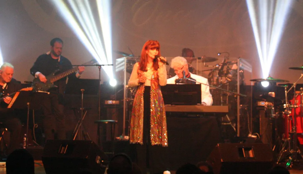 A band performs on stage with a female singer in a colorful outfit and musicians playing instruments highlighted by bright stage lights