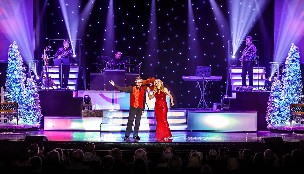 A festive stage performance features a man and woman singing under bright lights accompanied by musicians flanked by illuminated Christmas trees