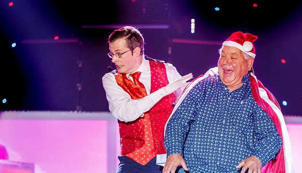 Two men are joyfully interacting on stage with one wearing a red festive hat and cape and the other in a red vest with a microphone
