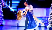 A man and woman in formal attire are performing a dance on stage, surrounded by Christmas trees and bright lights.