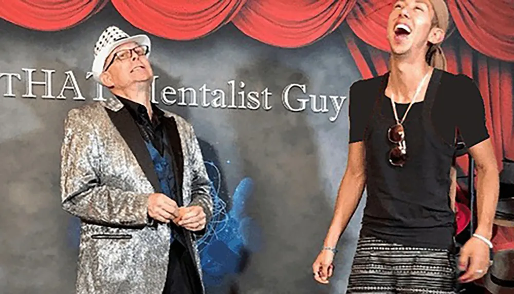 Two people are on stage one wearing a shiny jacket and hat and the other laughing joyfully with a backdrop that reads THA Mentalist Guy