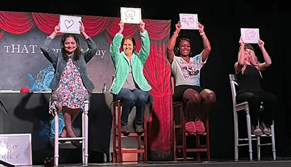 Four people seated on stage hold up sketches displaying them to the audience